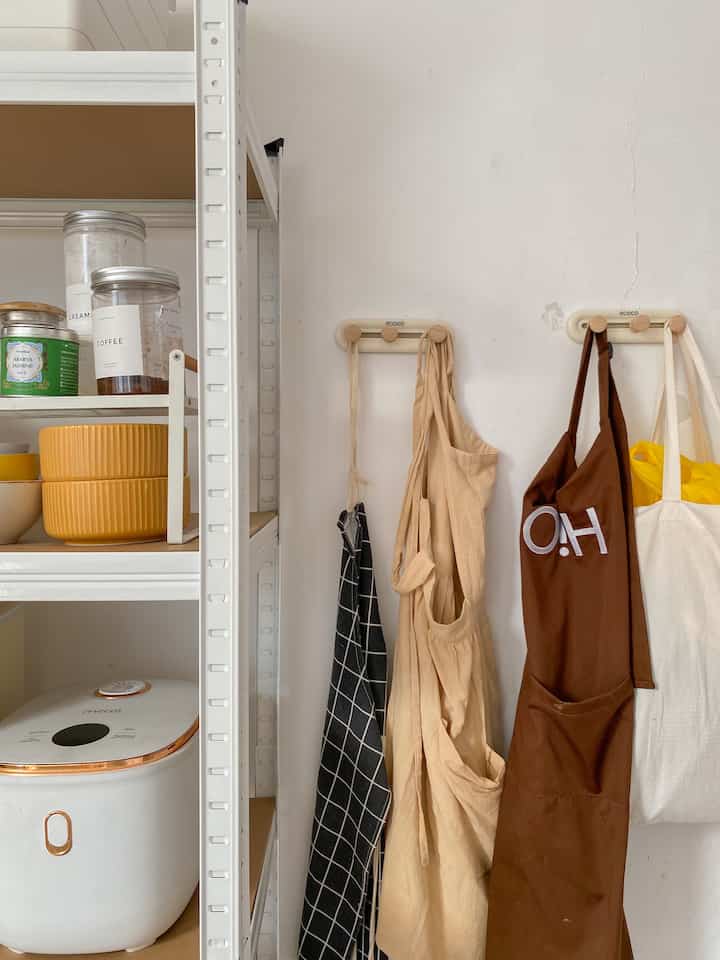 White and beige toned kitchen storage space featuring hanging aprons on wall hooks in a neat kitchen corner