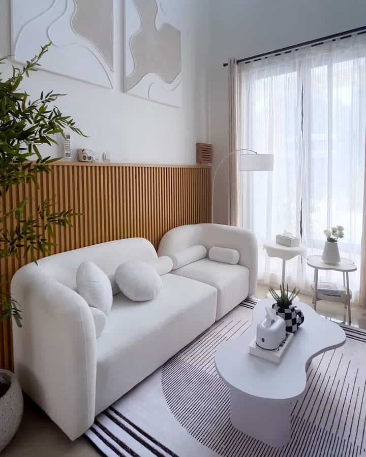 A white and natural wood tone living room featuring a simple sofa, coffee table, and sheer curtains creating a clean atmosphere