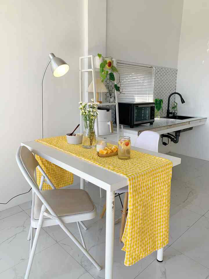 A compact kitchen with dominant white and yellow tones, featuring a white dining table, floor lamp, and window with blinds in a bright minimalist space