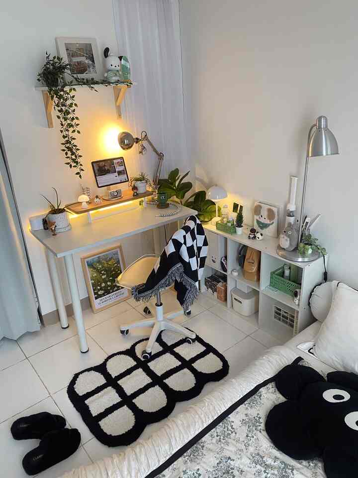 White-toned studio apartment featuring a central white desk with office chair, white sofa, and various plants creating a simple and practical interior