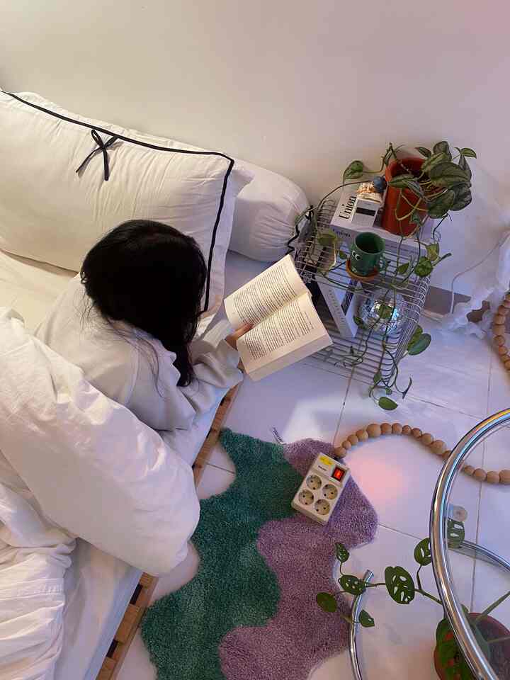 White and natural-toned living room featuring a person reading on a sofa bed next to a colorful carpet and plants, cozy atmosphere