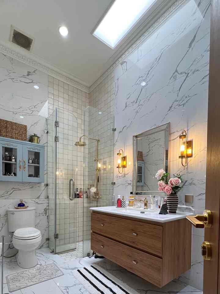 A modern bathroom in white and gold with marble tiled walls, transparent glass shower enclosure, and wood-toned vanity cabinet