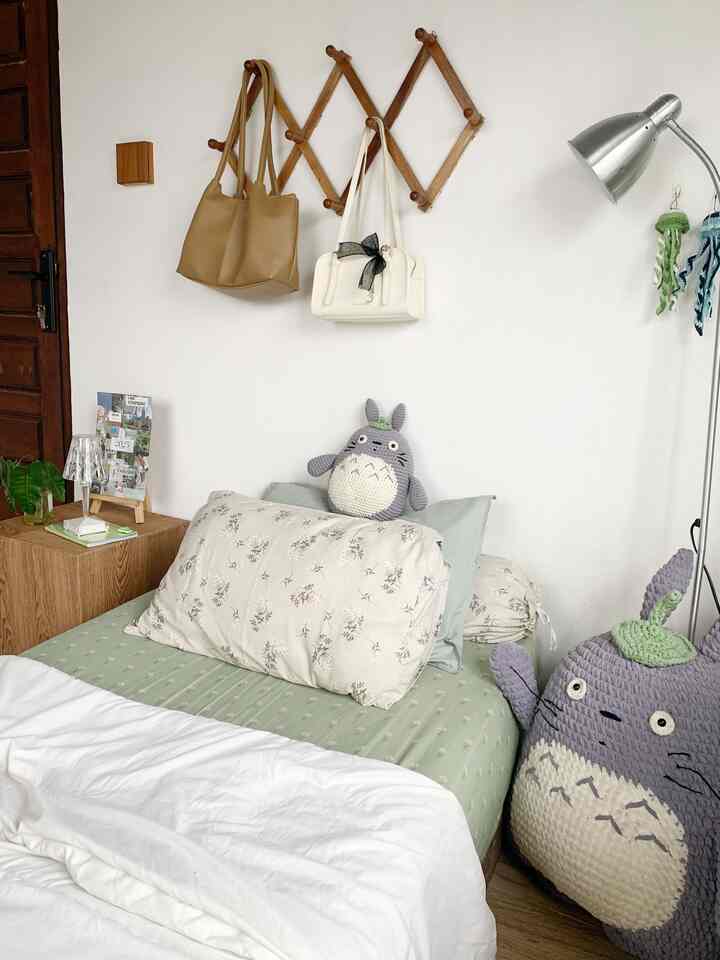 Bedroom with beige walls and green bedding, featuring Totoro plush dolls and wooden hooks, creating a cozy atmosphere