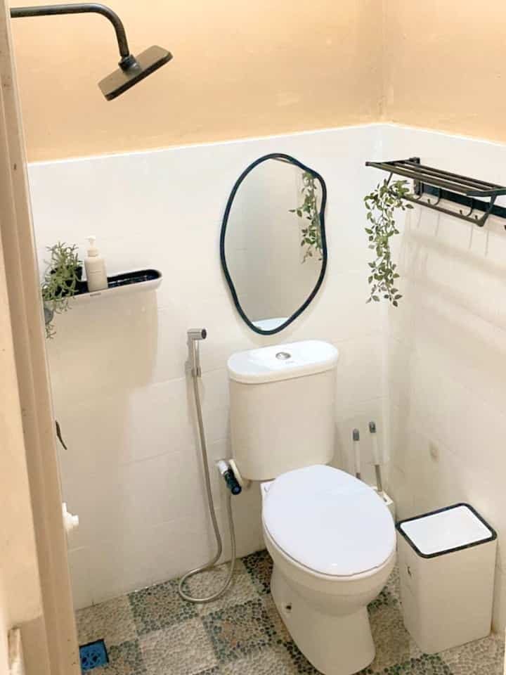 Small bathroom in beige and white tones featuring a toilet with bidet spray, black-framed mirror, shelves with plants, and minimal decor