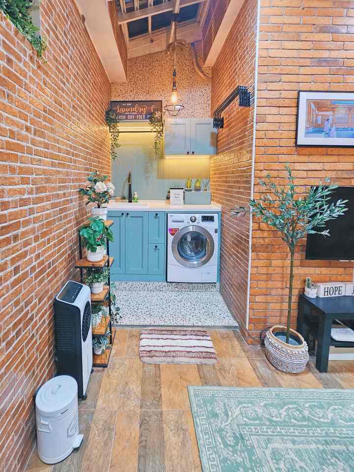 Natural style long, narrow room with orange-brown brick walls, blue kitchen cabinets, plants and washing machine arranged neatly