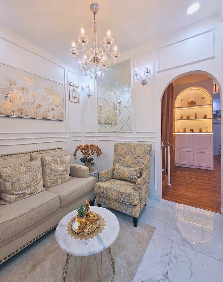 Beige and white toned living room featuring beige sofa, floral armchair, gold-accented coffee table, and crystal chandelier creating an elegant ambiance