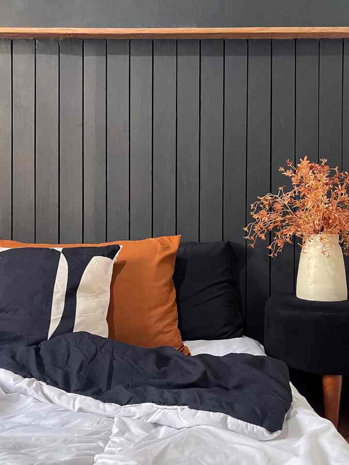 Bedroom with black paneled wall backdrop featuring brown cushions, a vase with dried flowers, and a black stool creating a calm atmosphere