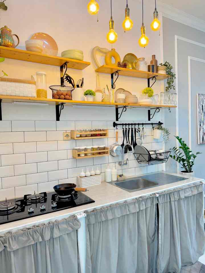 A warm kitchen featuring white tiled backsplash, wooden shelves with dishes, yellow filament pendant lights, and gray curtains covering lower cabinets with sink.