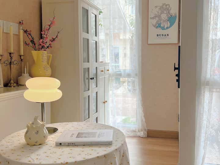 A cozy cafe-style small living room in beige and white tones featuring a round table, table lamp, and lace curtains with a warm atmosphere