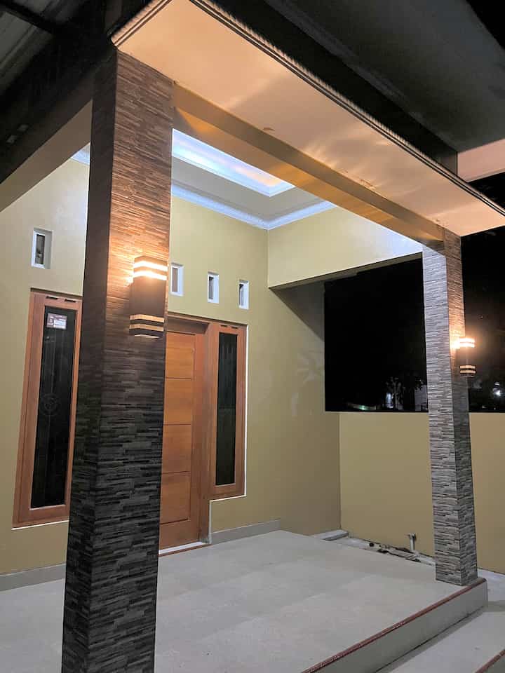 Modern balcony entry porch featuring yellow walls, brown wooden door, stone pillars with warm outdoor wall sconces lighting