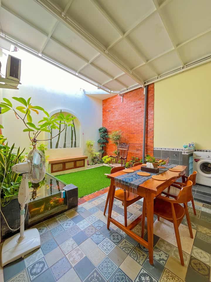 Natural wood tones and green plants harmonize in a semi-outdoor dining area with orange brick wall and patterned tile floor accents