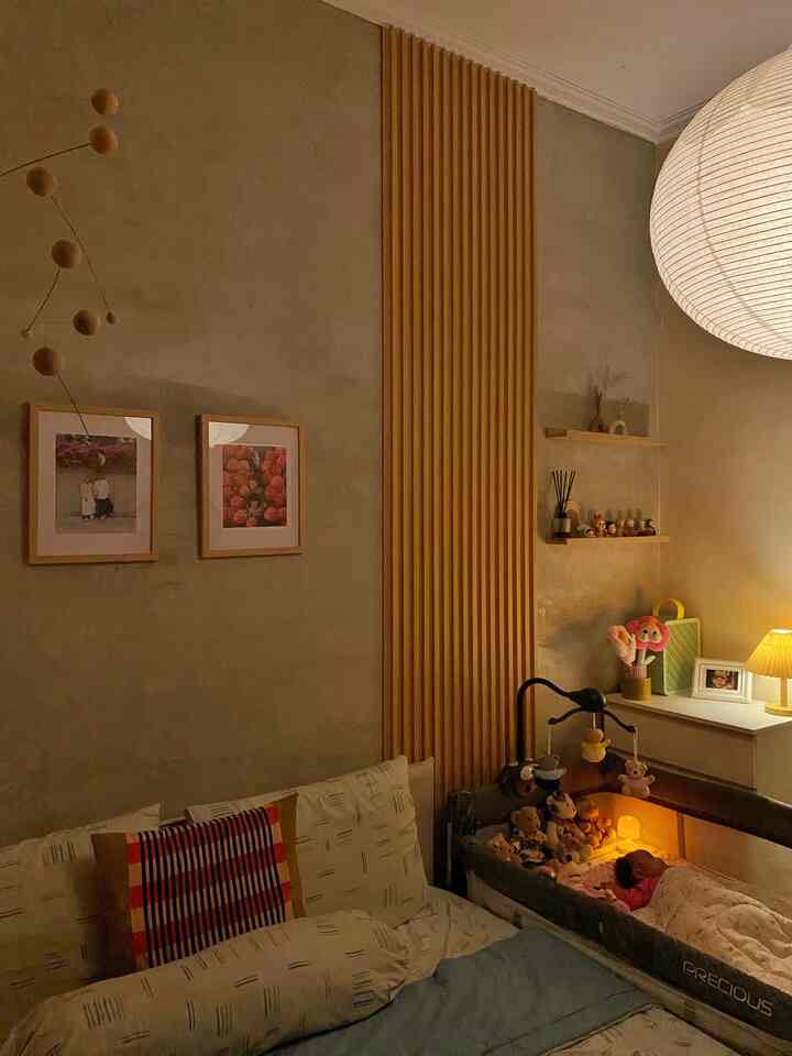 Warm beige toned bedroom featuring a bed, baby crib with mobile, and a table lamp creating a cozy atmosphere