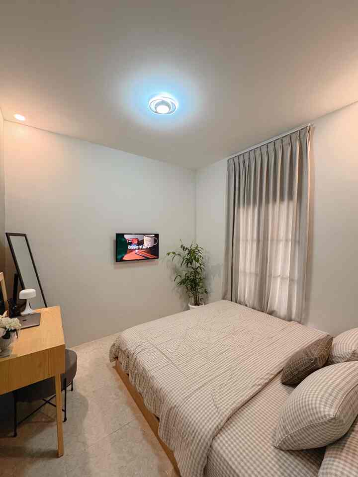 Bright natural-tone bedroom featuring a checked bedspread, beige curtains, and a wooden desk in a simple layout