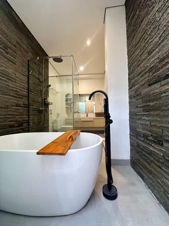 Natural toned bathroom featuring a large white bathtub with wooden tray, transparent glass shower enclosure, and textured stone walls creating a luxurious atmosphere