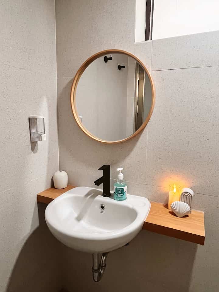 A bathroom with white walls and wood tone shelf featuring a round mirror, wash basin, and a lit candle creating a cozy atmosphere