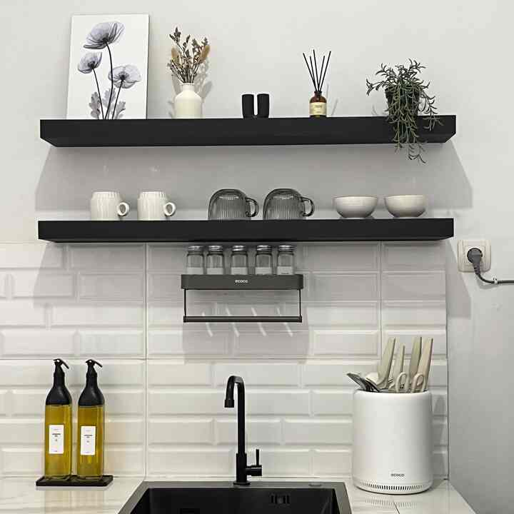 Clean kitchen space with white tiled wall, black shelves, a sink area, various kitchen utensils, and a plant for natural decor