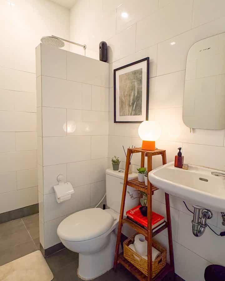 Compact bathroom with white tiled walls, dark gray floor, wood tone shelf, toilet, and sink arranged neatly