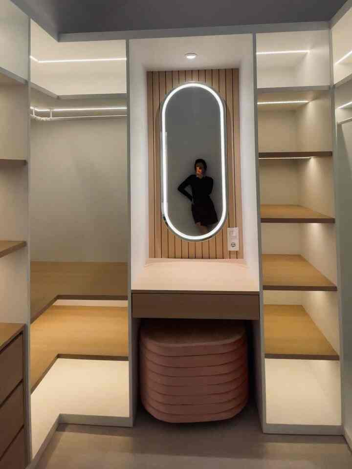 Natural wood-toned, simple, storage-focused compact walk-in closet featuring a central lit mirror with a cushioned stool