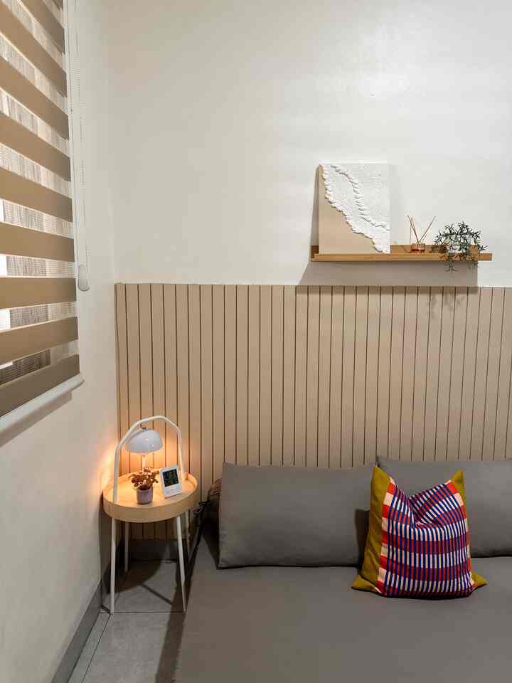 A simple, natural living room with beige wood tones, featuring a side table with lamp and a colorful cushion centerpiece