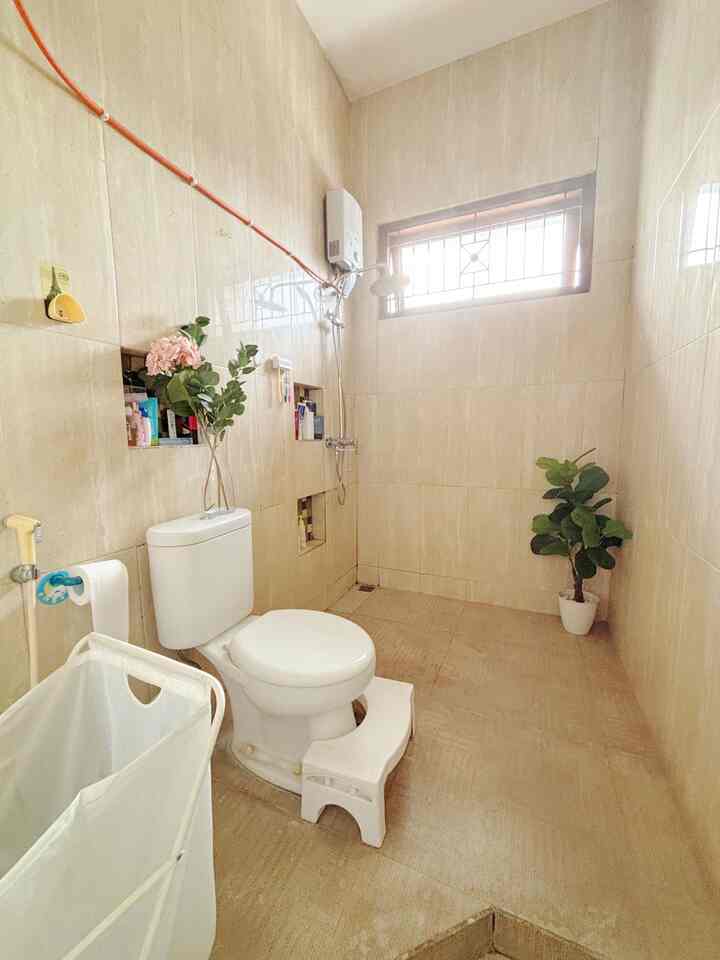 Beige-tiled bathroom featuring a white toilet, step stool, and plants in a simple, clean setting with natural light