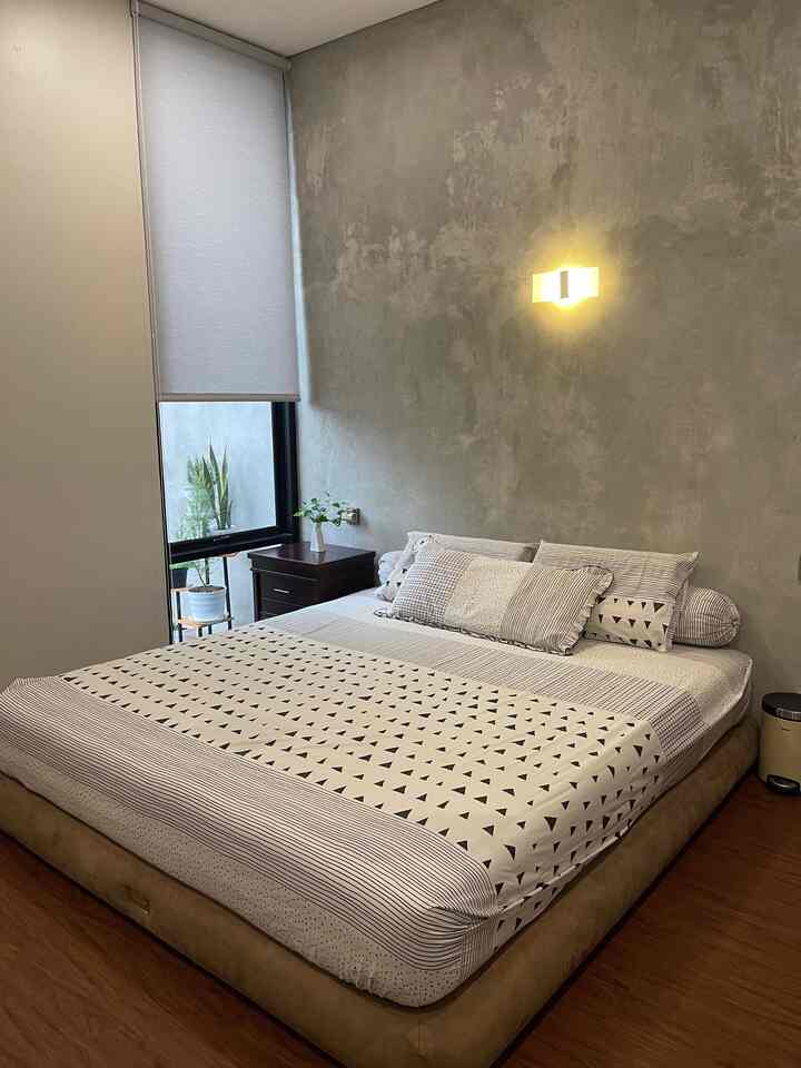 Natural modern tone bedroom featuring beige bed, exposed concrete wall, white bedding, and wall sconce lighting