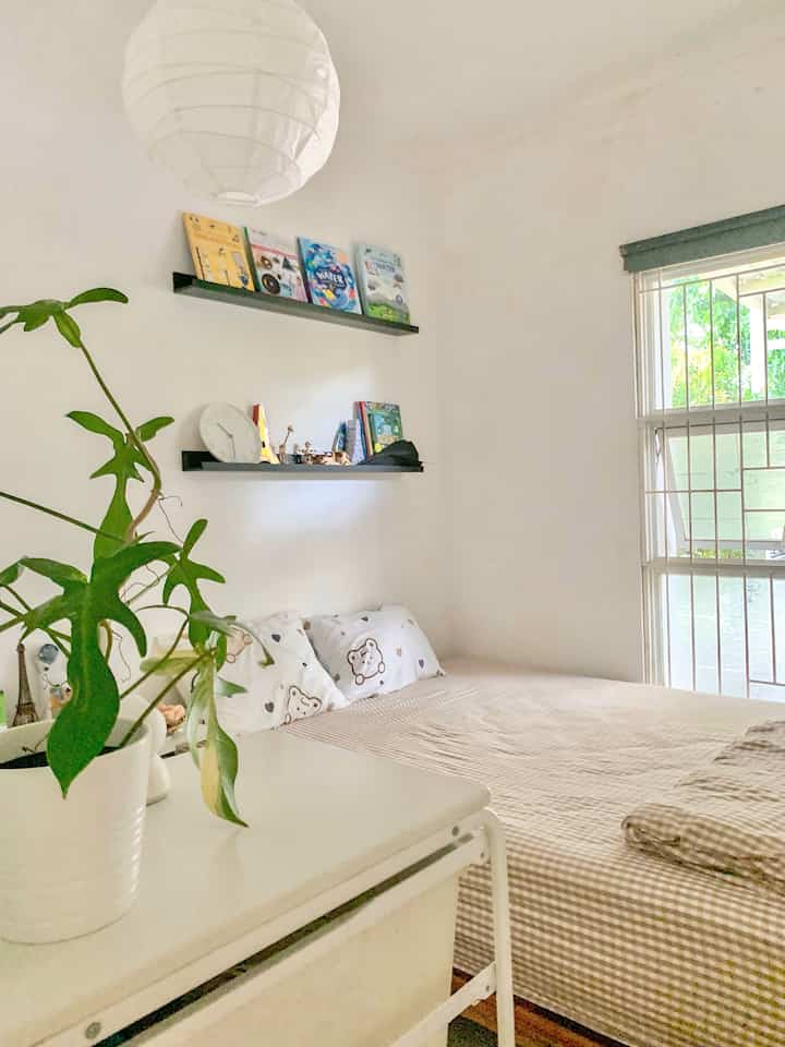 A white-walled kids' bedroom featuring beige bedding, minimalist shelves with books, and a simple blackout curtain creating a cozy atmosphere