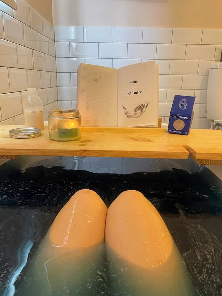 Bathroom with white tiles and wooden tray, featuring a marble bathtub with partially submerged legs for a relaxing bath