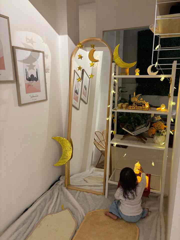 White-walled small living room with rugs, a baby near a full-length mirror adorned with moon and star ornaments, warm yellow lighting creating cozy atmosphere