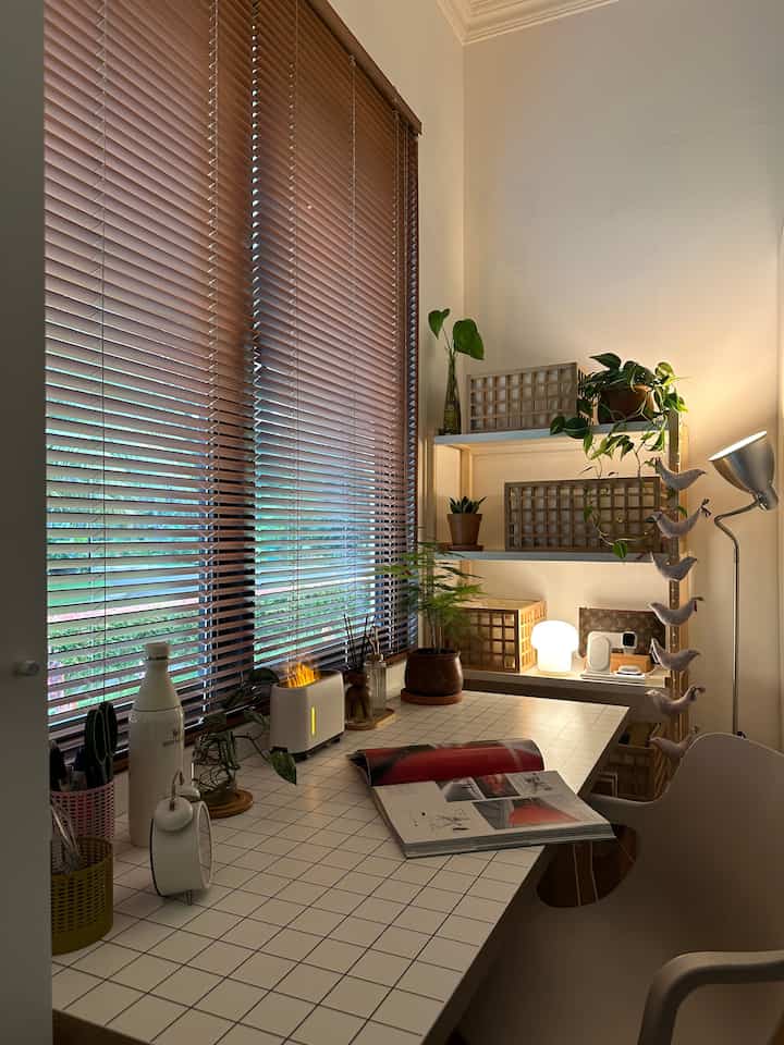 Cozy home office featuring wood tone blinds and a white desk, adorned with plants adding a natural touch