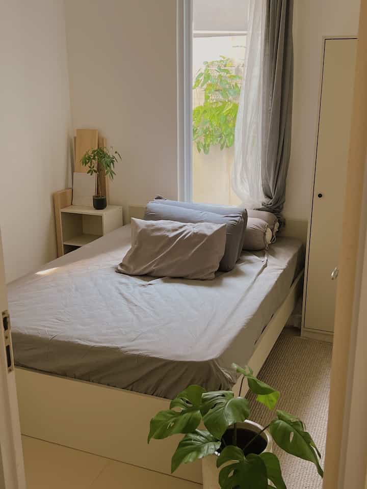 A small, minimal bedroom in natural gray and beige tones featuring a bed, curtains, and indoor plants for a cozy atmosphere