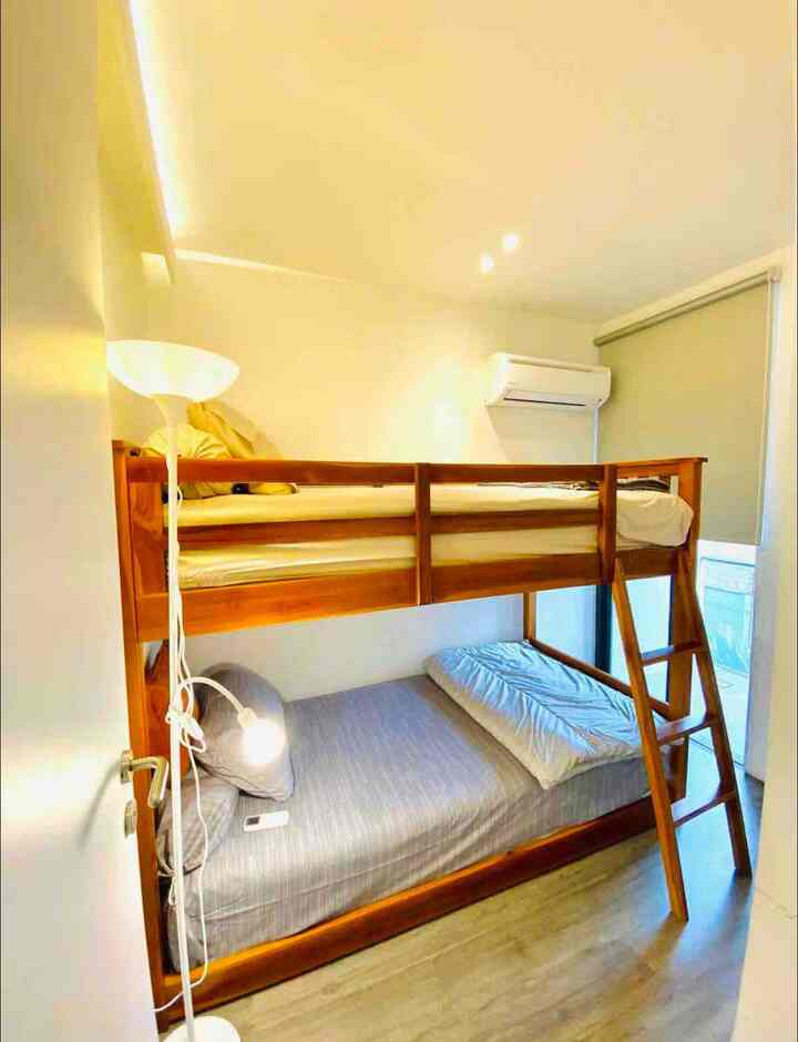 White-walled compact bedroom featuring a wood-toned bunk bed, a floor lamp near the door, and a roller blind covering the window for a cozy atmosphere