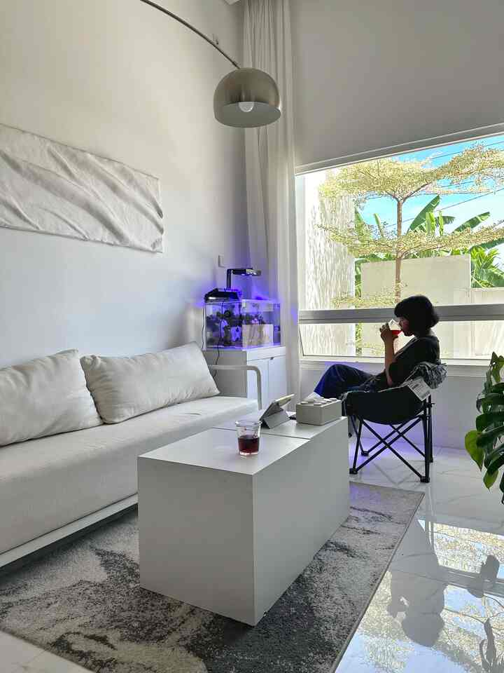 White and gray toned living room featuring a white sofa, coffee table, large window, and a folding chair by the window in a modern clean space