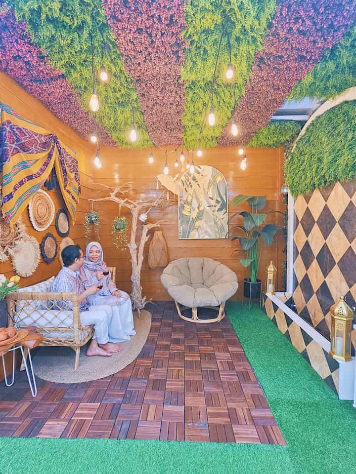 Green and wood-toned balcony space featuring rattan sofa, cushioned chair, plants and lighting for a cozy cafe-style interior
