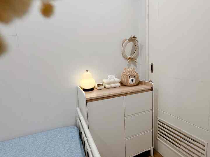 White-toned compact small room featuring a table lamp and baby-themed storage cabinet with soft decor items
