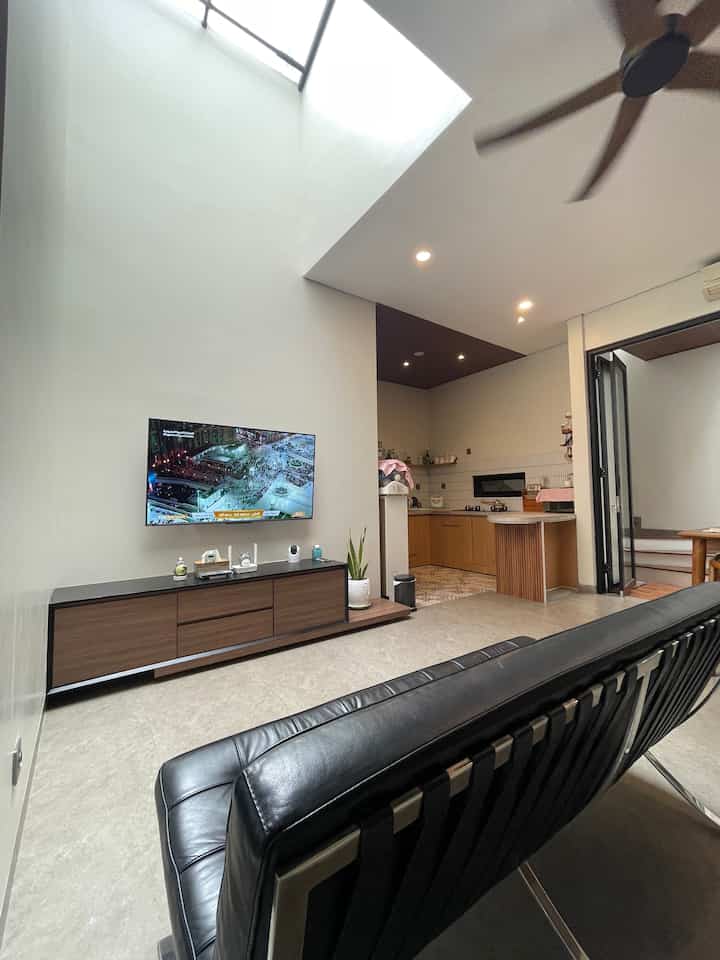 Modern living room and kitchen with white walls and wood tone furniture, featuring a black leather sofa centrally placed
