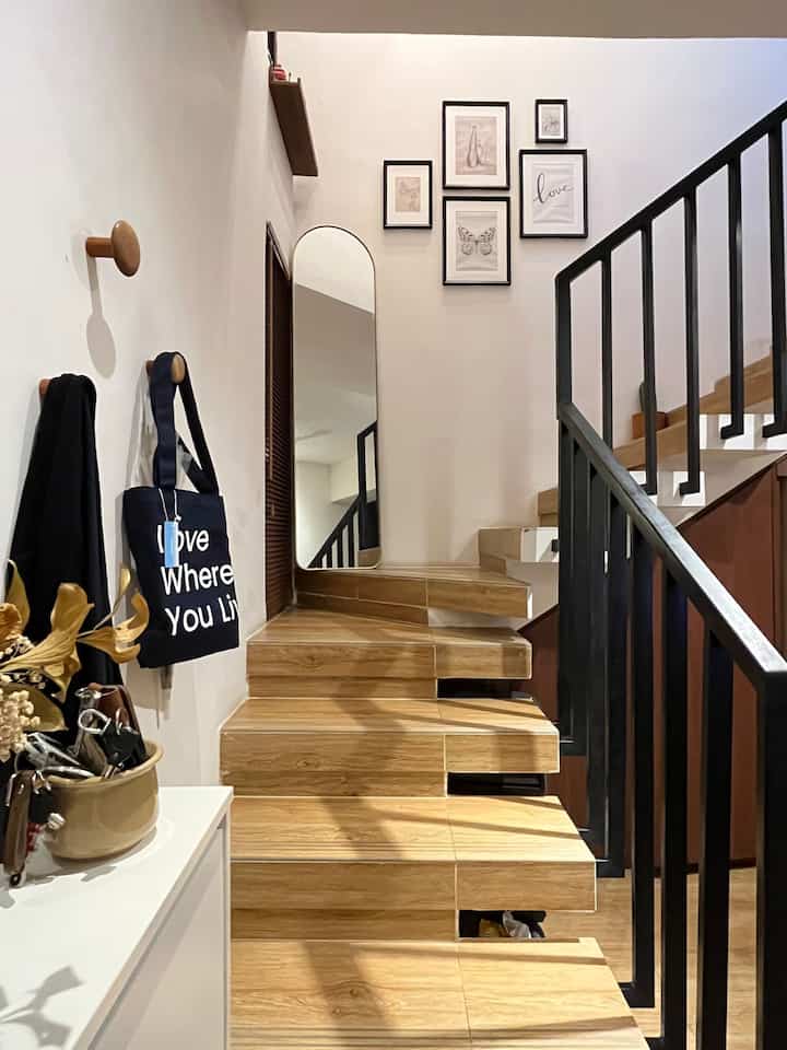 Modern entrance space with wood-toned stairs and black railing, featuring framed art and a tall mirror on the wall