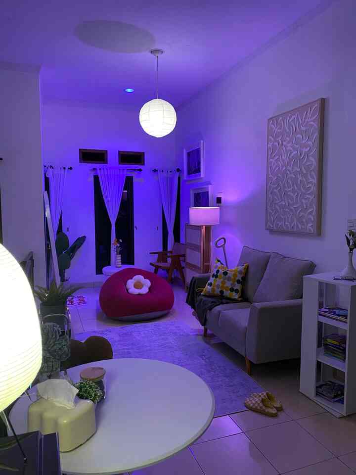 White-toned living room softly lit by purple lighting, featuring a gray sofa and pink bean bag creating a cozy atmosphere