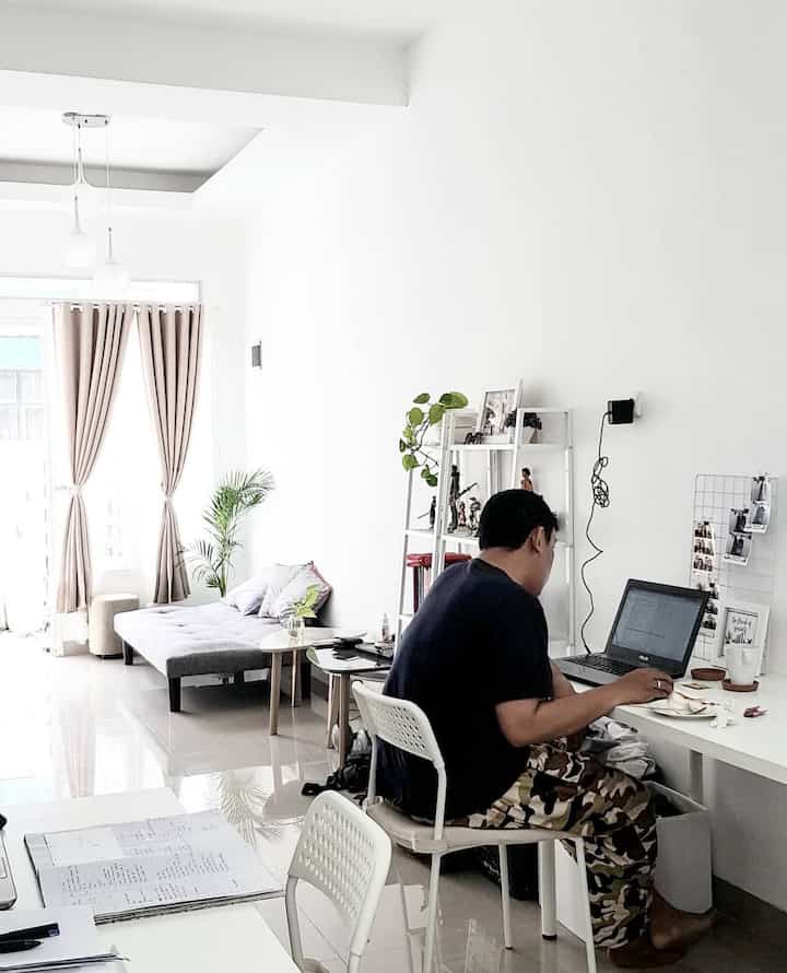 Bright white and beige compact home office featuring a sofa bed, desk, bookshelf, and plant in a modern interior