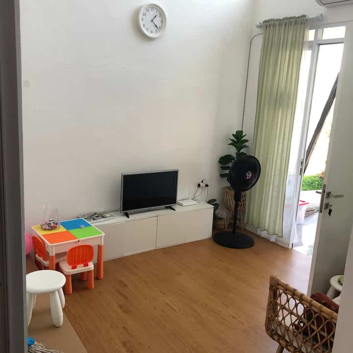 Natural toned living room with wood tone flooring, white TV stand and soft green curtains in a compact-sized space
