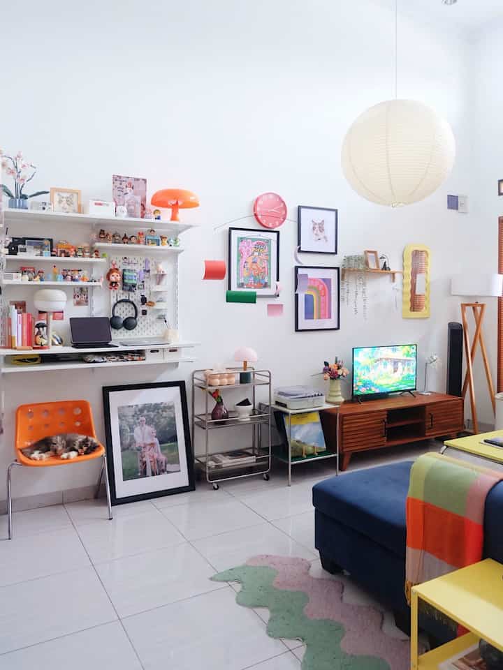 White-walled living room in mid-century modern style featuring an orange chair and lamp, blue sofa, and colorful carpet creating a bright, cheerful space.