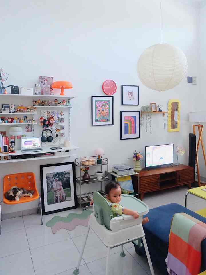 White-based living room with orange accents, featuring a baby in high chair and a pet cat on an orange chair amidst colorful decor