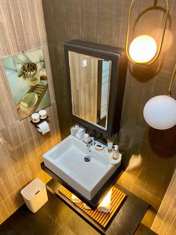 Modern bathroom with brown tiled walls and floor, white sink with a large mirror, wooden shelf, and wall sconces