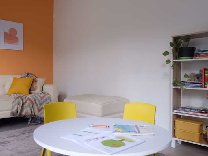 Bright and simple kids' room with white and orange accent walls, featuring yellow chairs, a white round table, and a bookshelf