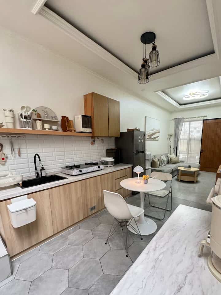 Natural-toned and white open space kitchen and living room featuring wood cabinetry, modern lighting, and neat minimalist design