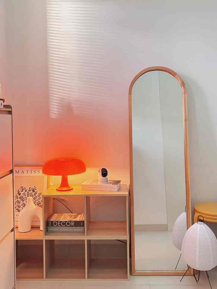 Cozy beige-toned living space featuring an orange lamp, wooden standing mirror, and shelving unit with decorative books and items.
