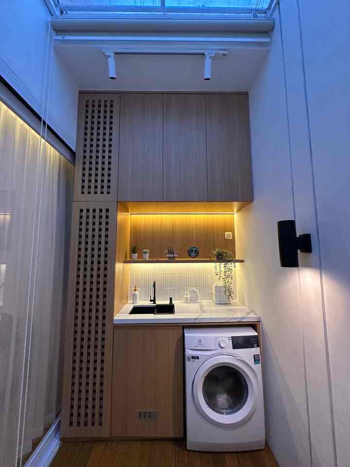 Minimal narrow laundry room with white walls and wood tone cabinets, featuring a washing machine, sink, and natural light from a skylight