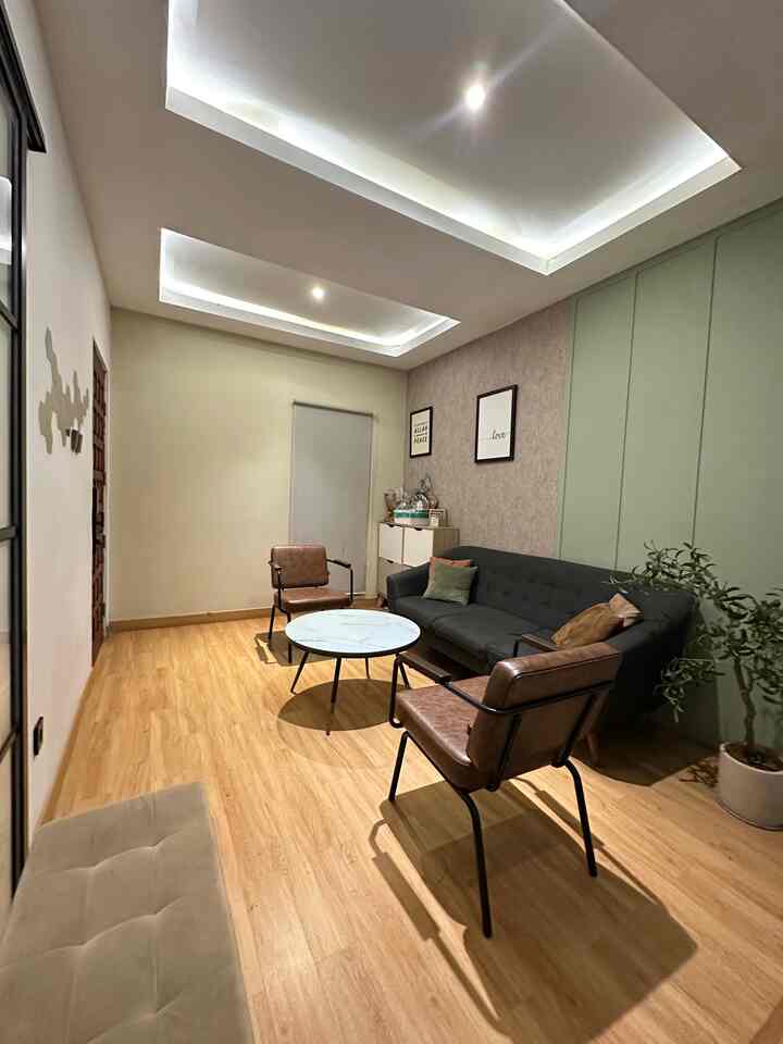 Minimalist living room with natural brown wood flooring and green accent wall, featuring simple sofa and armchairs creating a cozy atmosphere