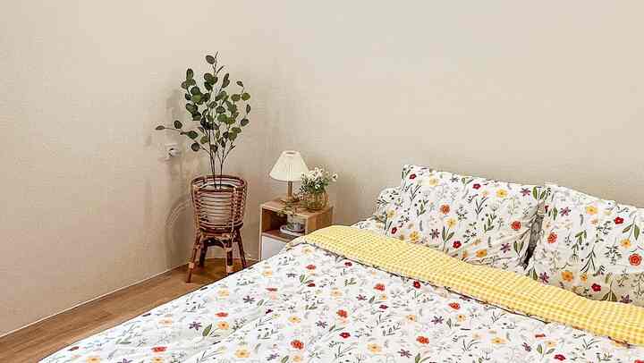 Beige-walled bedroom with wood tone flooring featuring floral bedding and a rattan plant stand creating a natural atmosphere