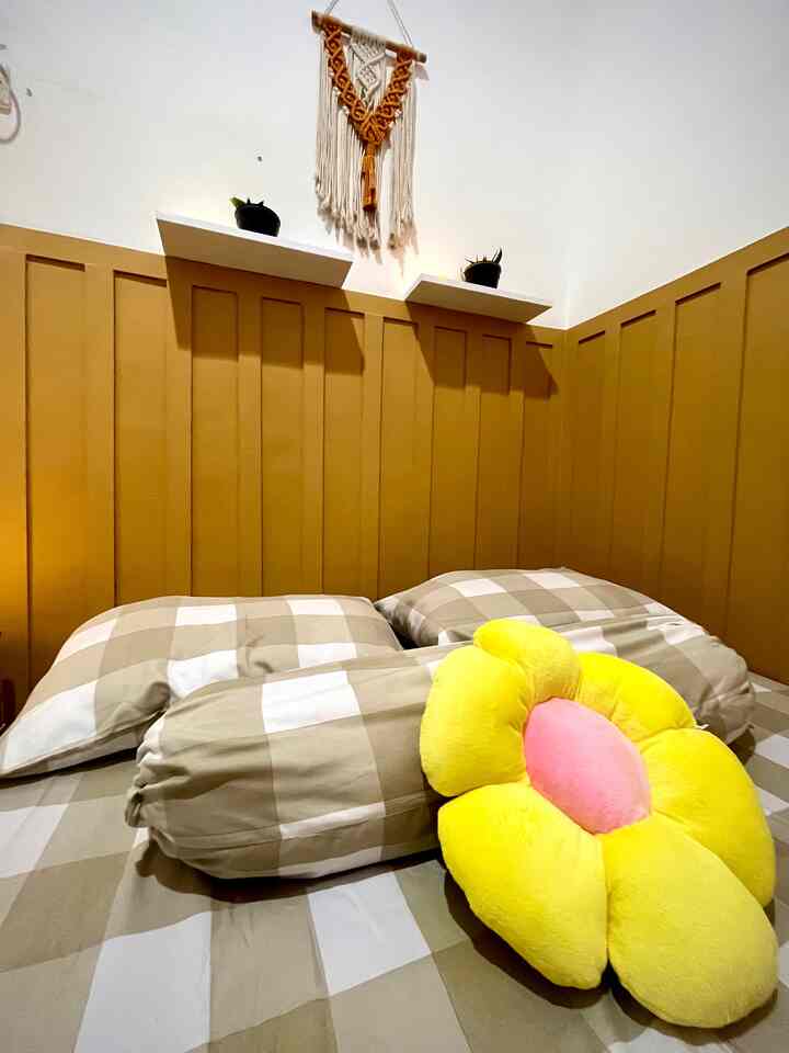 A cozy, simple bedroom with mustard yellow paneled walls and beige checkered bedding accented by a bright flower-shaped cushion