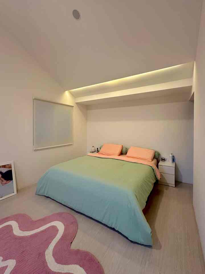 Simple bedroom in white and beige tones featuring a centrally placed bed with bedding and a pink rug, creating a cozy atmosphere